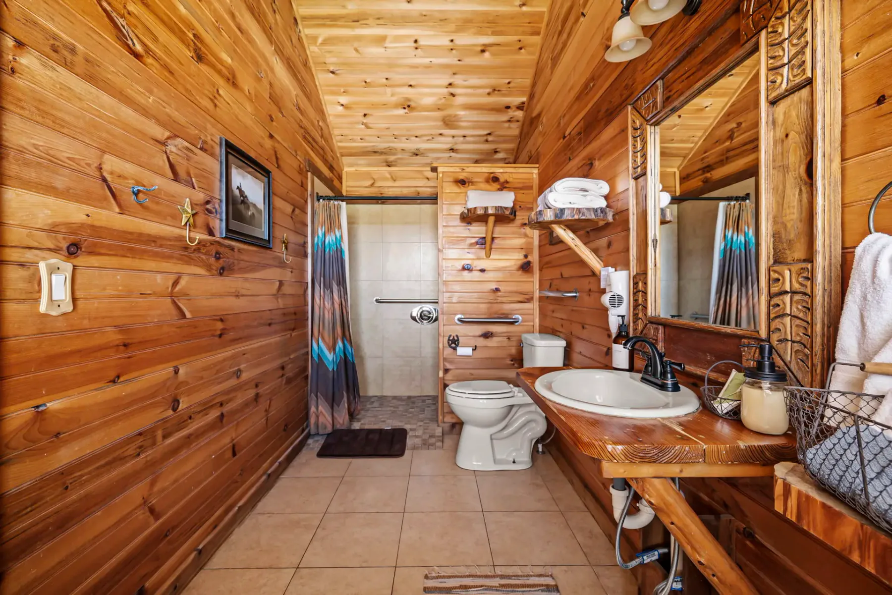Baño rústico de madera con piso de baldosas, ducha, inodoro, lavabo pequeño, espejo, toallero y diversos accesorios.