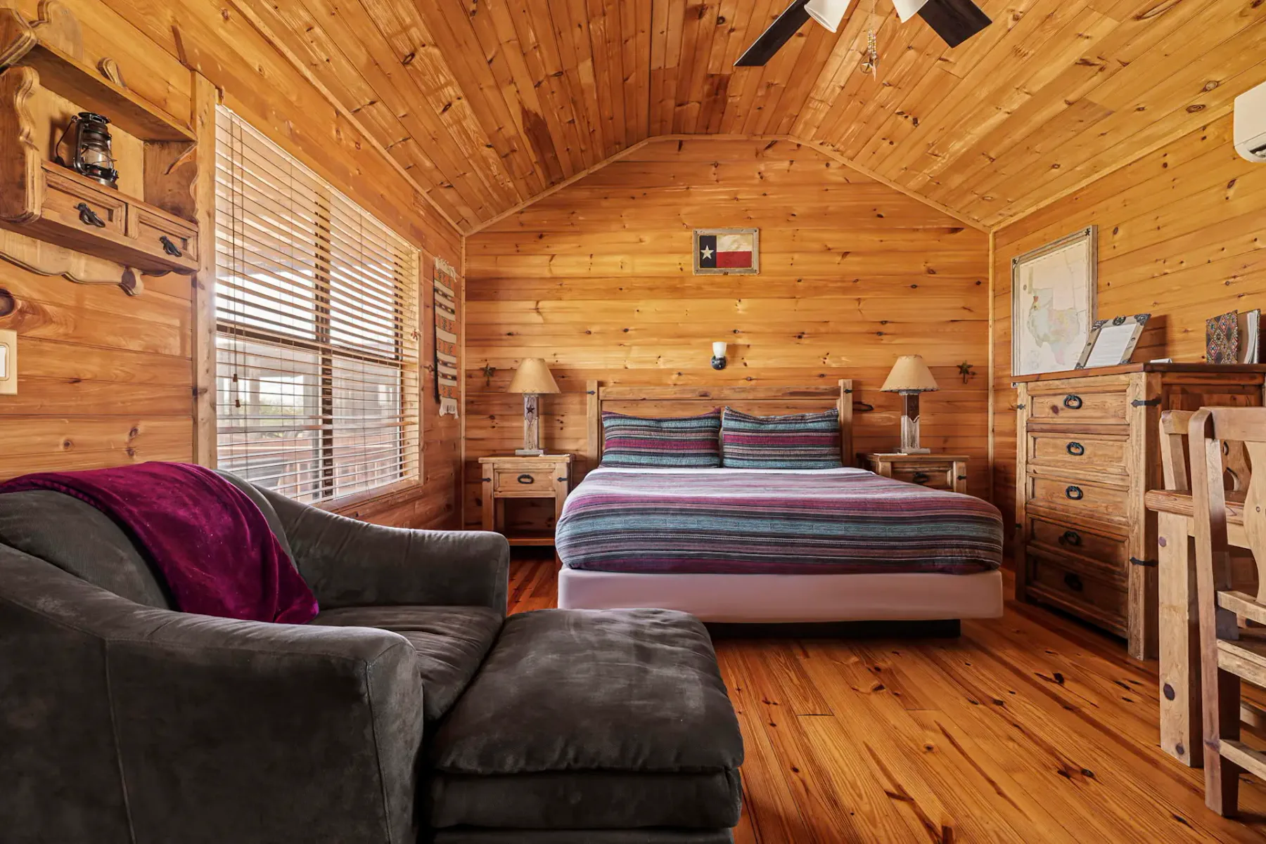 Acogedor dormitorio de cabaña de madera con una cama con ropa de rayas, muebles rústicos, sofá gris e iluminación suave procedente de una ventana y un ventilador de techo.