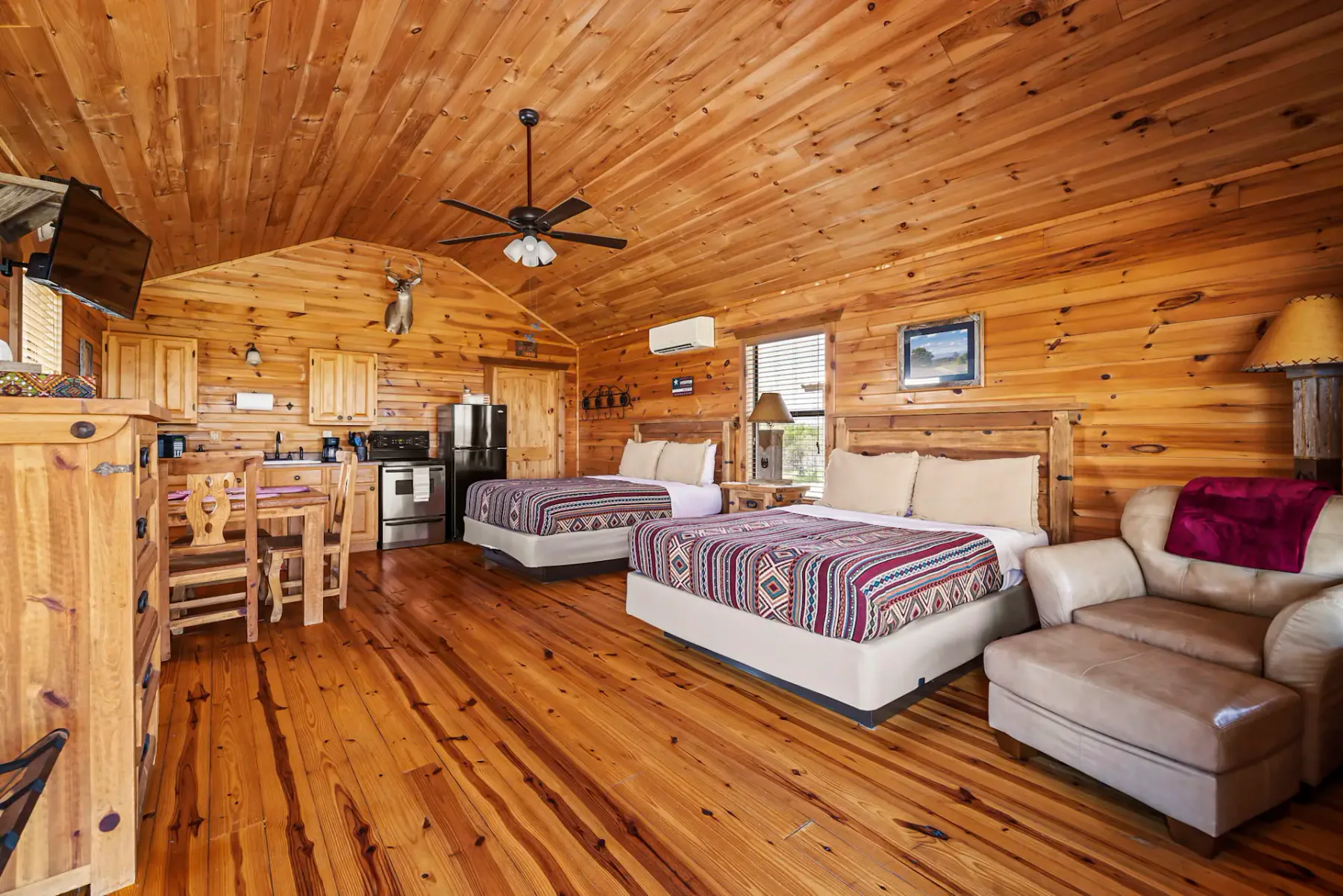 Interior acogedor de una cabaña de troncos con dos camas, un pequeño comedor y electrodomésticos modernos de cocina, con paredes y pisos de madera de tonos cálidos.