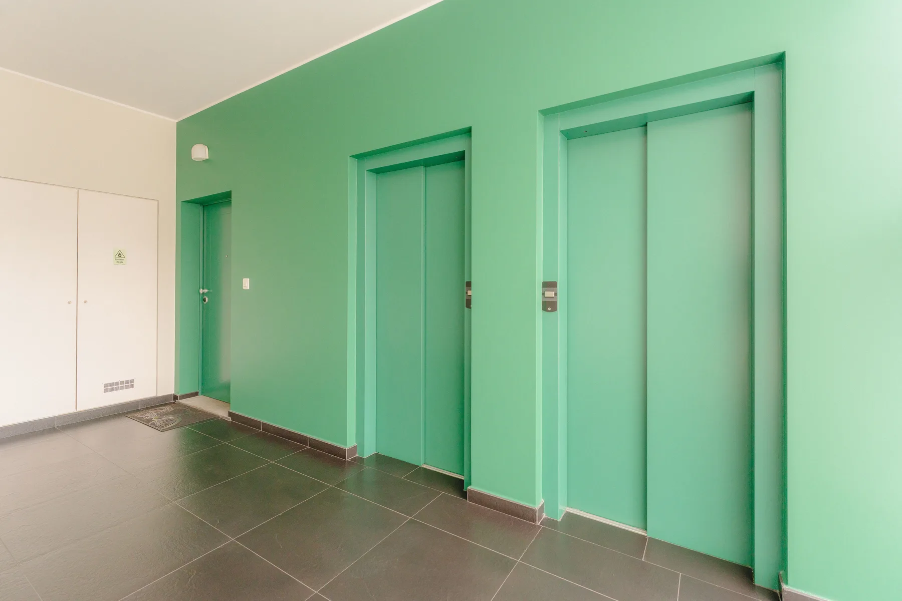 Un vestíbulo de ascensores verde menta con varias puertas de ascensor y suelo de baldosas oscuras.