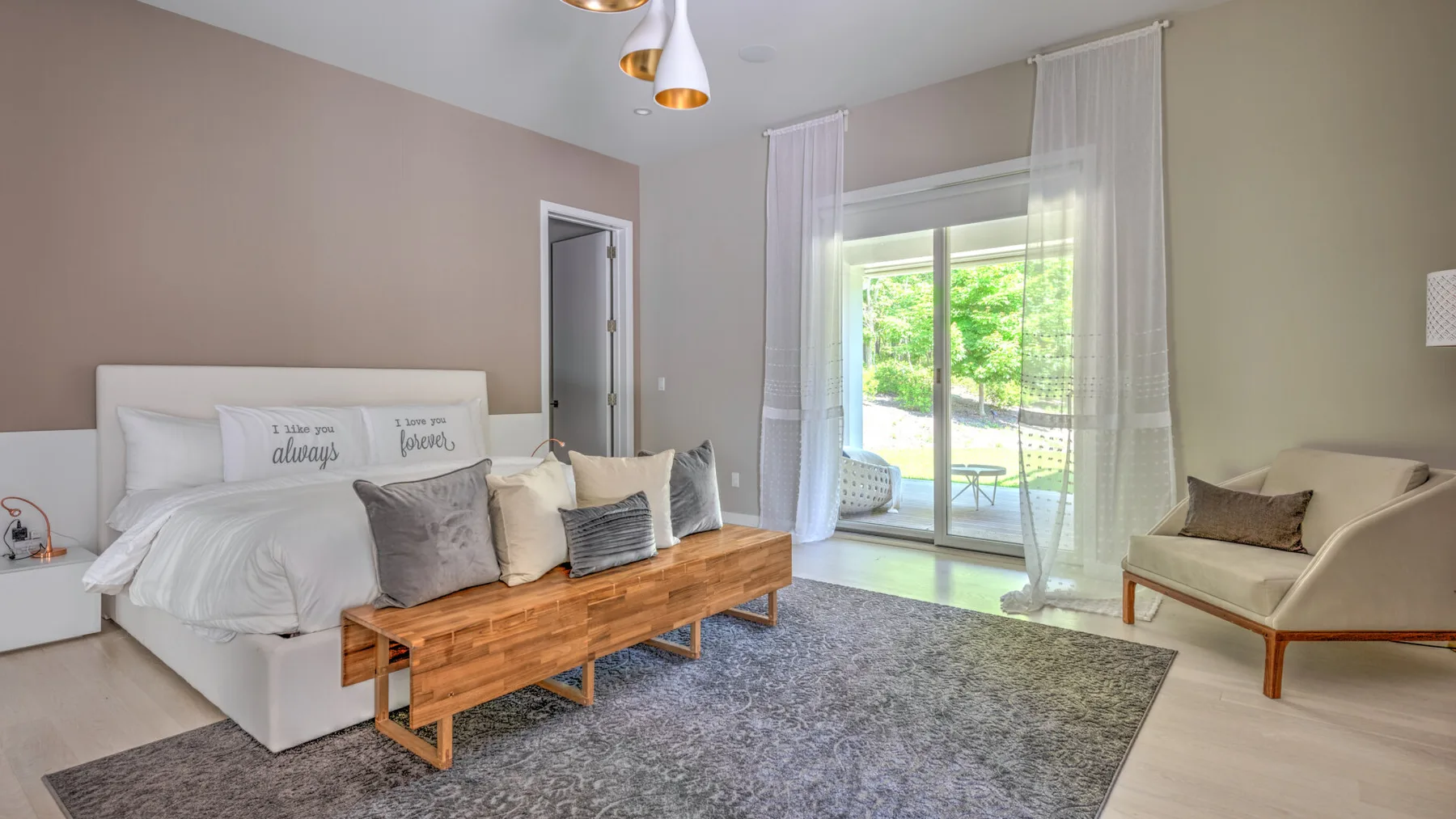 Un dormitorio luminoso con cama, banco de madera y grandes ventanas con vistas a la vegetación.