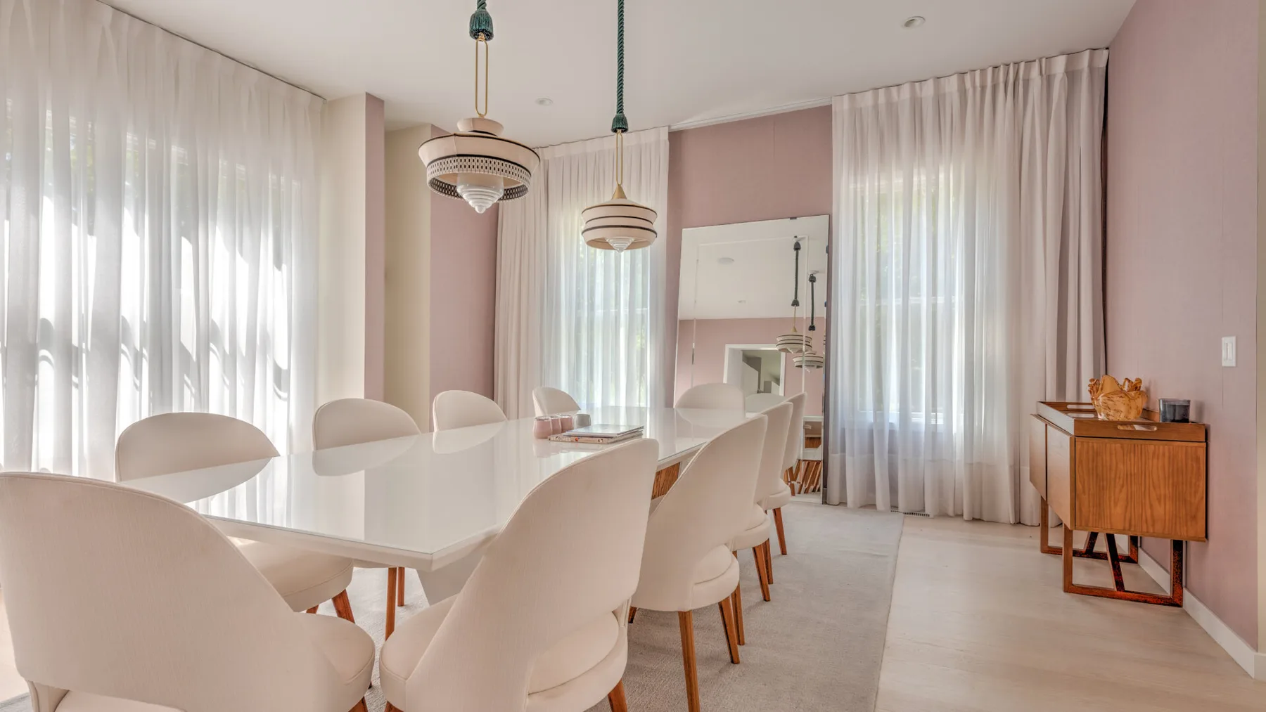 Un comedor con una mesa blanca alargada, asientos tapizados y cortinas altas.