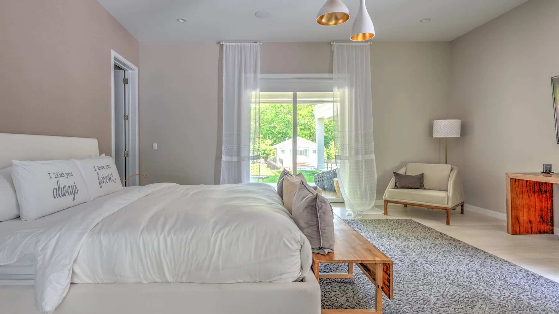 Un dormitorio luminoso con una cama junto a cortinas translúcidas y una pequeña zona de estar.