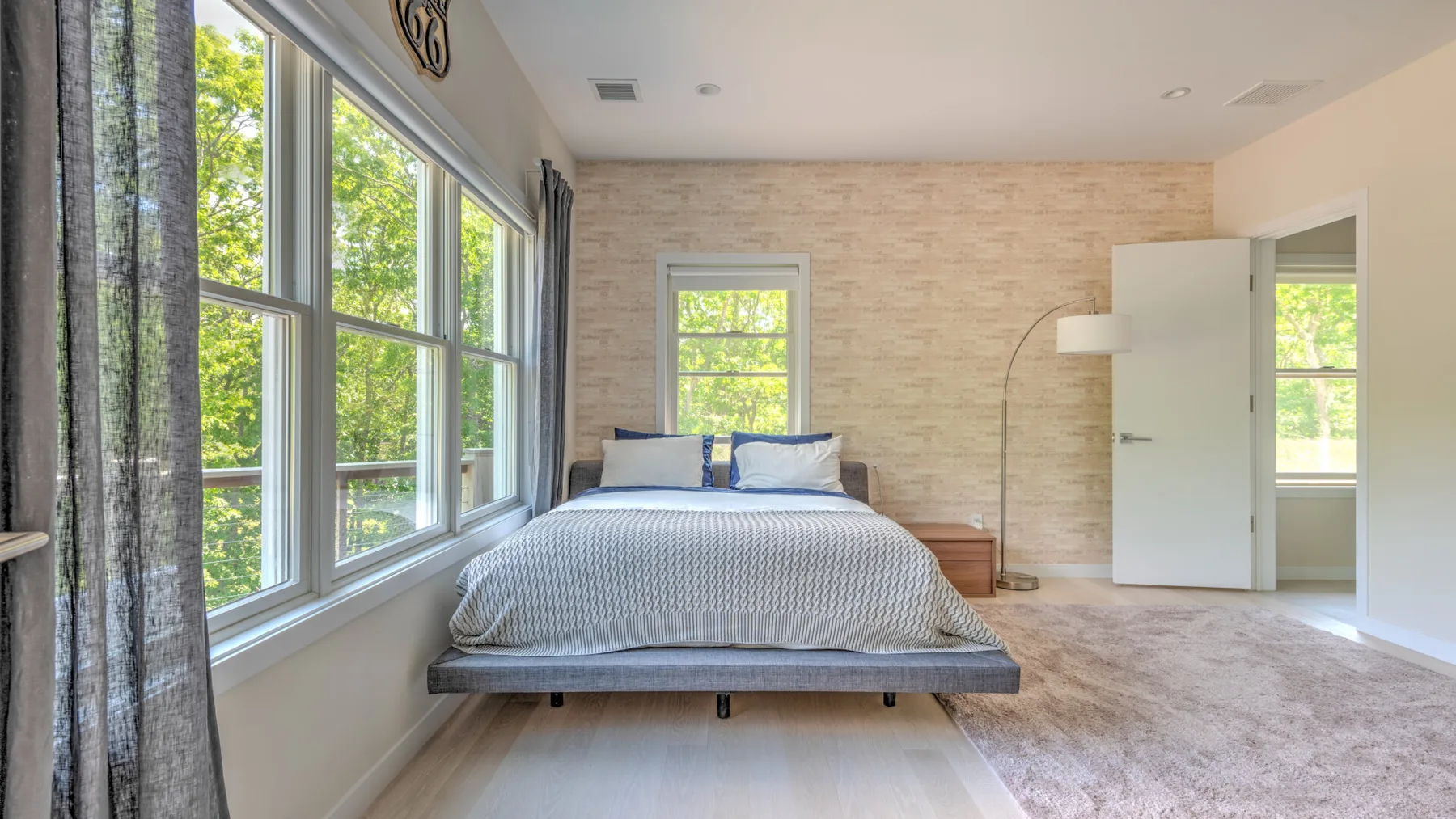 Un dormitorio luminoso con una cama junto a grandes ventanas y una pared texturizada.