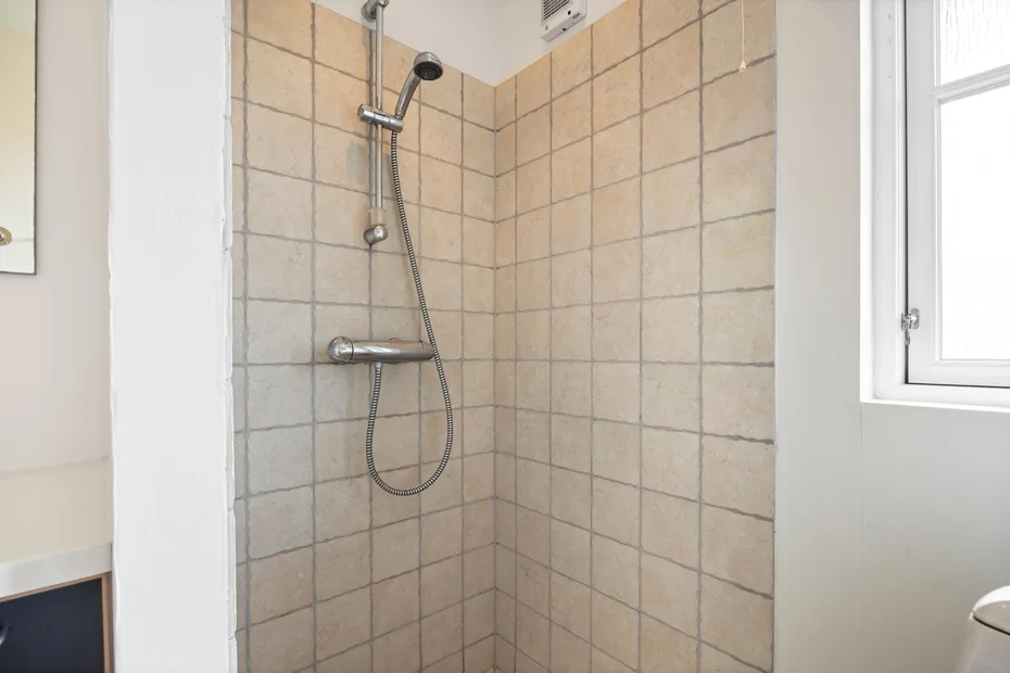 Una zona de ducha con azulejos, una ducha de mano y una ventana cercana.