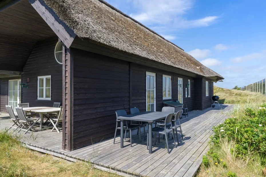 Una casa de madera oscura tiene una amplia terraza con asientos junto a dunas cubiertas de hierba.