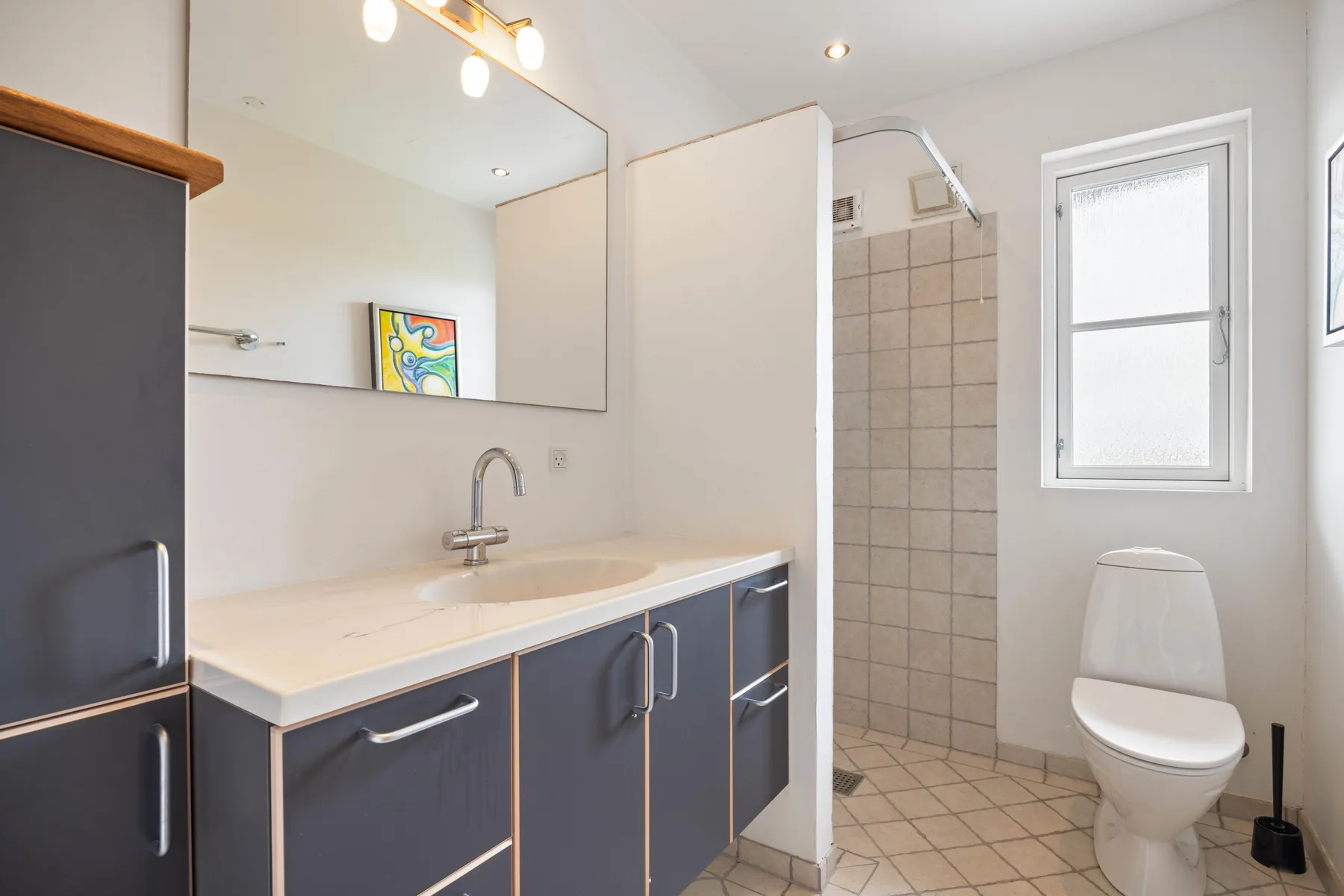 Un baño luminoso con un tocador largo, una ducha con azulejos y un inodoro.