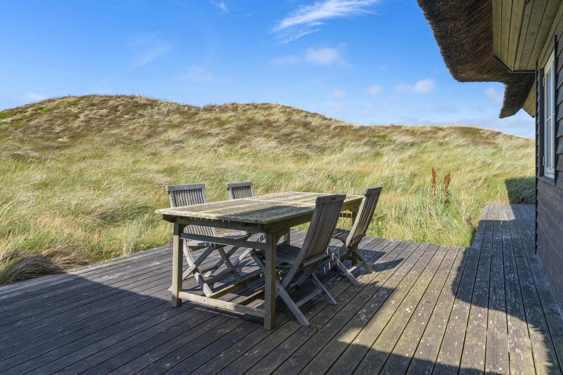 Una terraza exterior con mesa y asientos junto a dunas cubiertas de hierba.