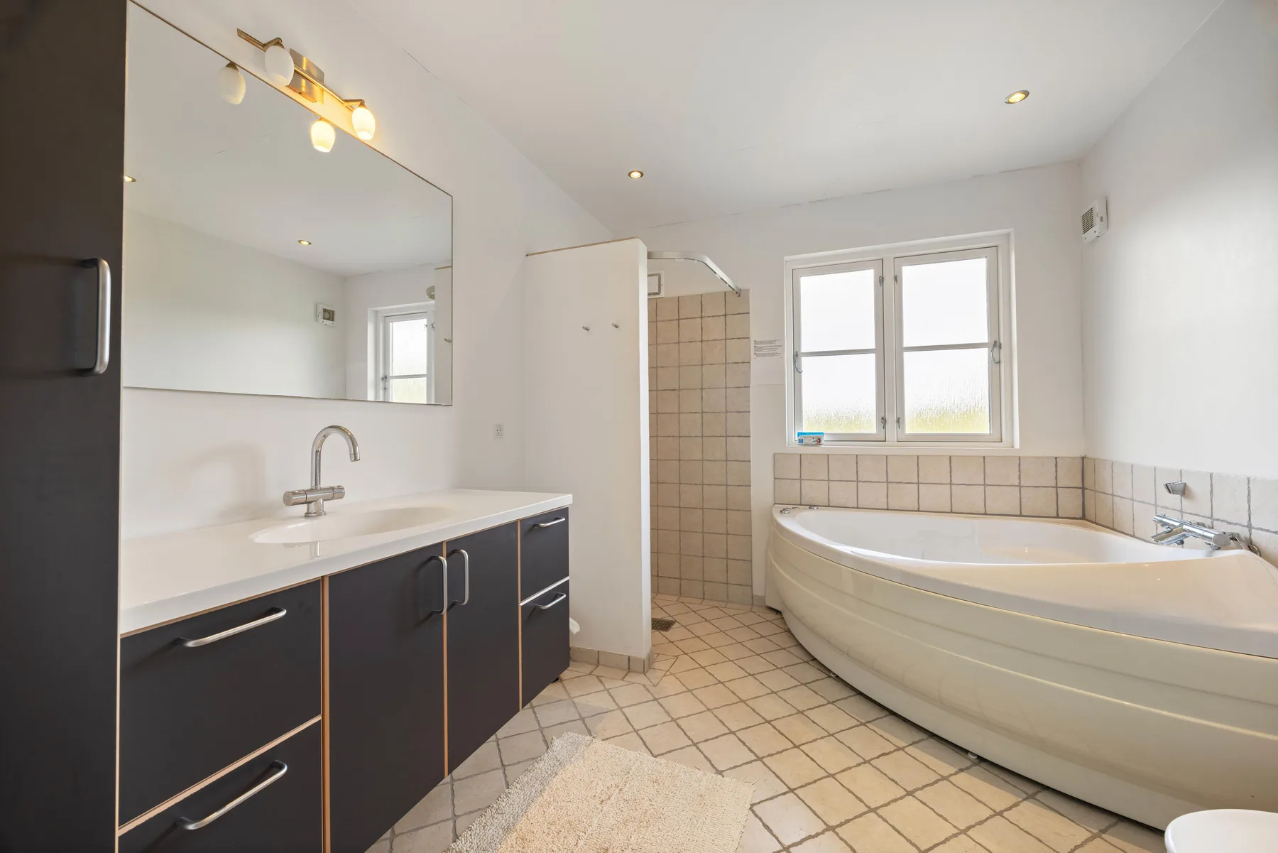 Un baño luminoso tiene un tocador, una ducha con azulejos y una bañera bajo dos ventanas.