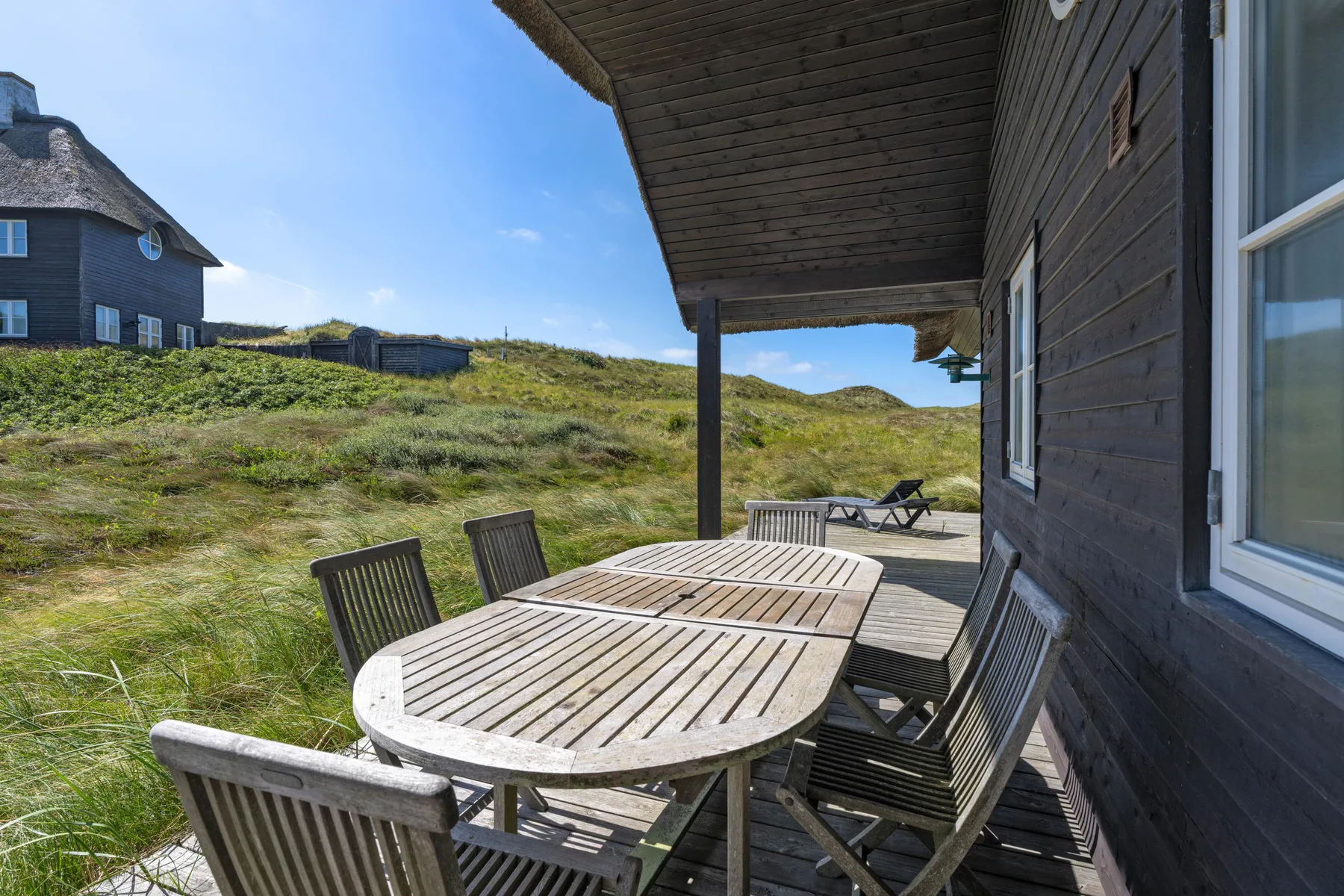 Una zona de comedor exterior de madera junto a una casa da a dunas cubiertas de hierba.