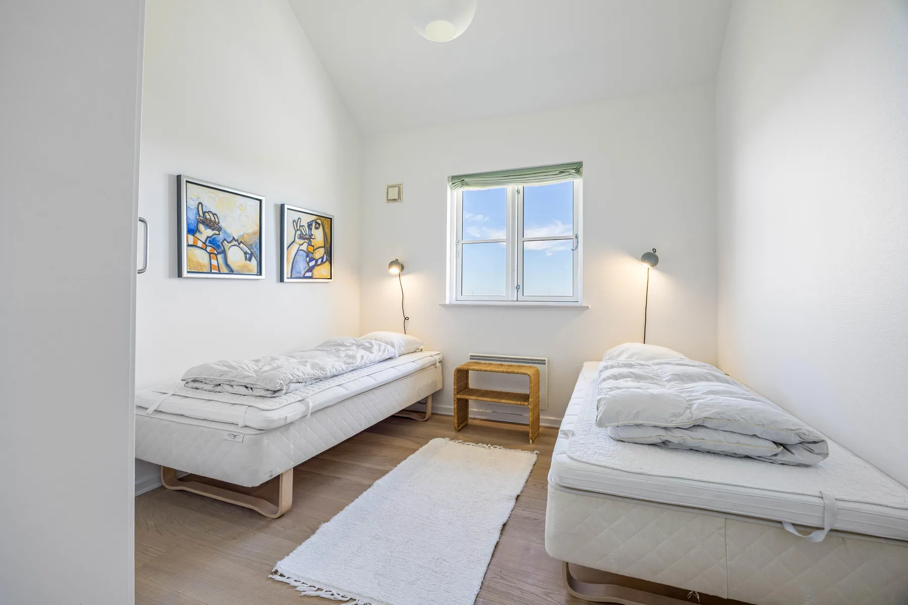 Un dormitorio luminoso con dos camas, una ventana y cuadros enmarcados.