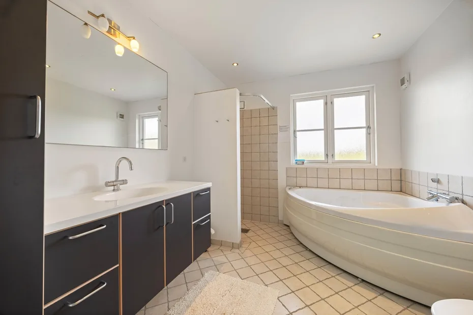 Baño luminoso con un largo tocador, zona de ducha con azulejos y bañera esquinera.