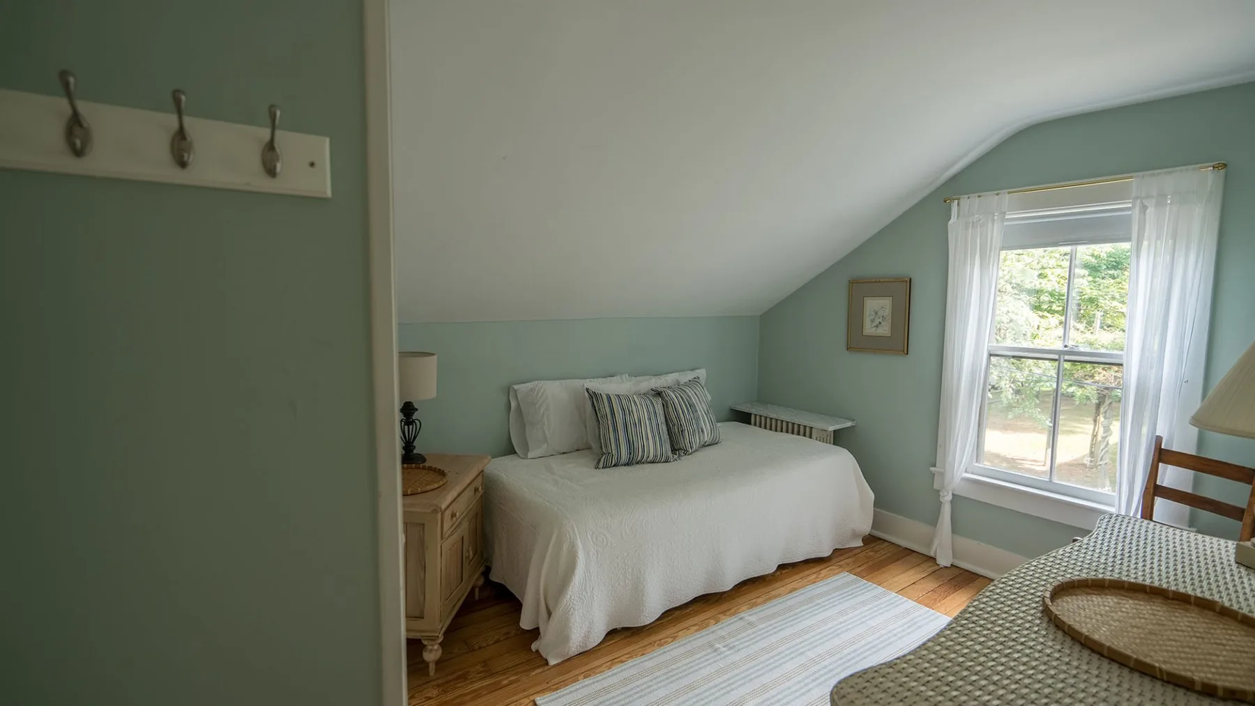 Un dormitorio con una cama bajo un techo inclinado y una ventana con cortinas transparentes.