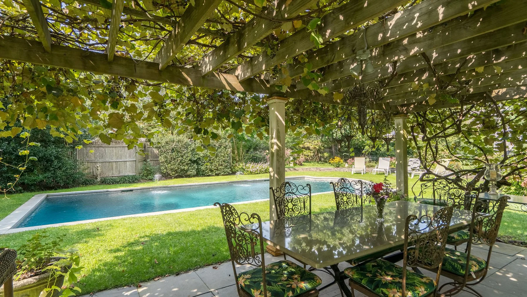 Una zona de comedor al aire libre con sombra da a un césped y una piscina.