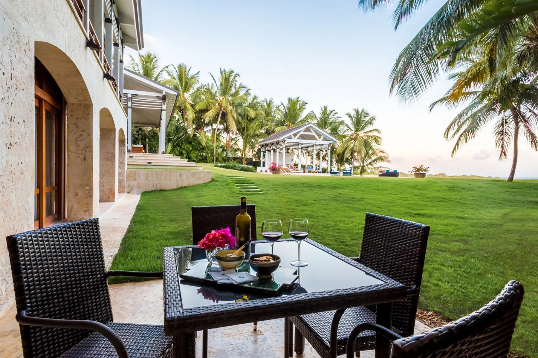 Una zona de comedor al aire libre da a un césped, palmeras y un pabellón blanco.
