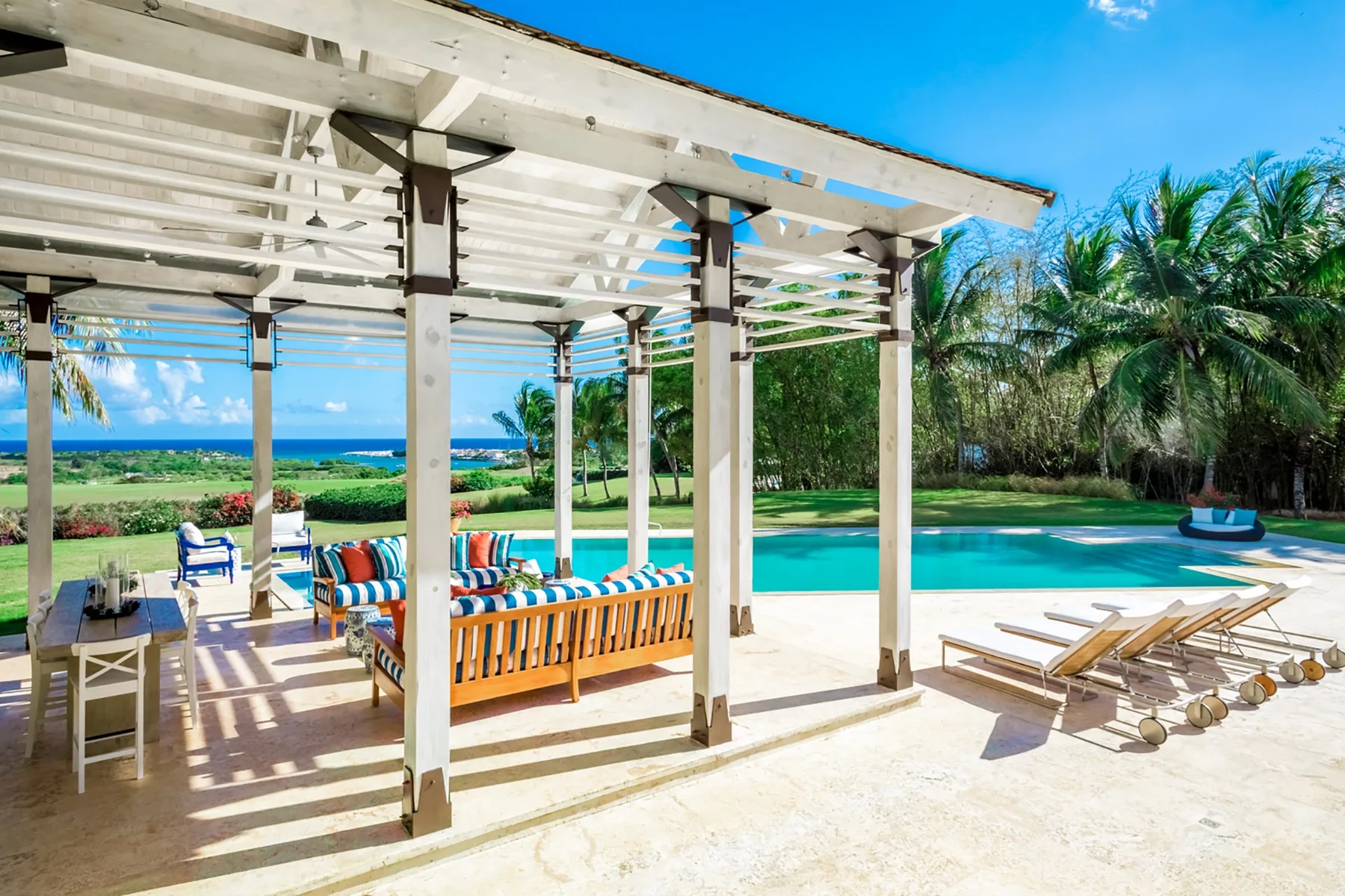 Una zona de estar al aire libre con sombra da a una piscina y palmeras.