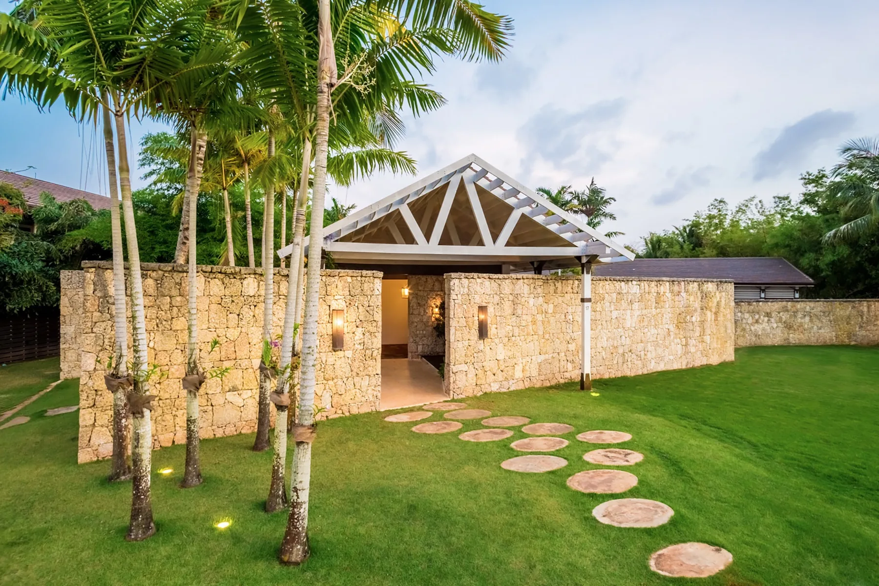 A stone building stands on a lawn beside palm trees and a circular stepping-stone path.