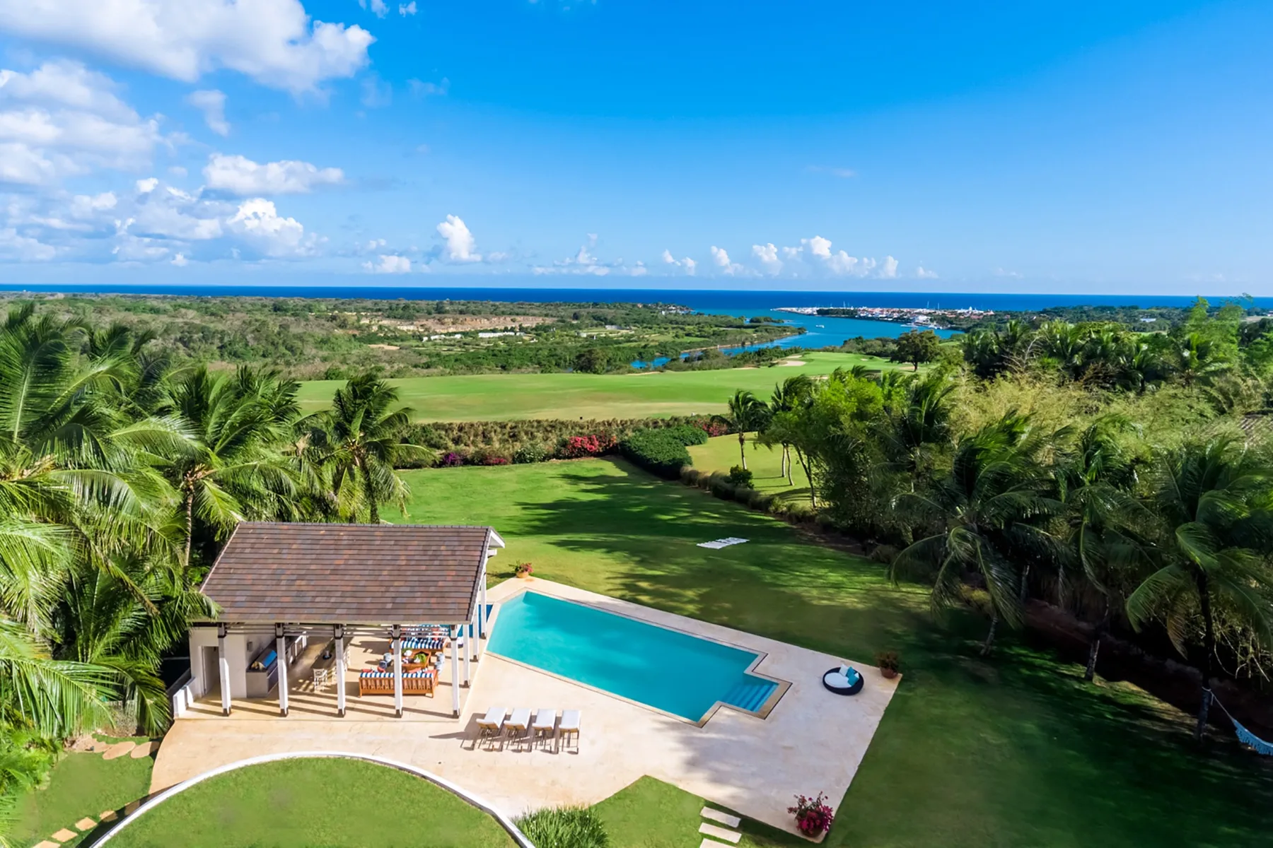 Aerial view of a swimming pool and pavilion amid palm trees, with the ocean in the distance.