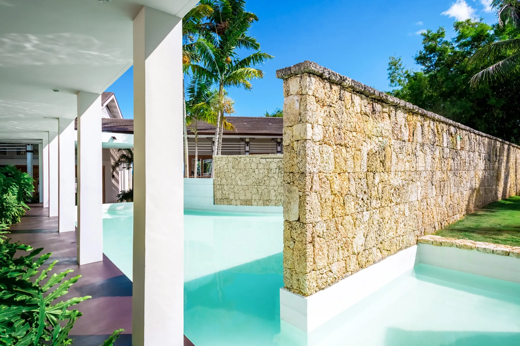 Una piscina turquesa junto a un alto muro de piedra y un pasillo cubierto.