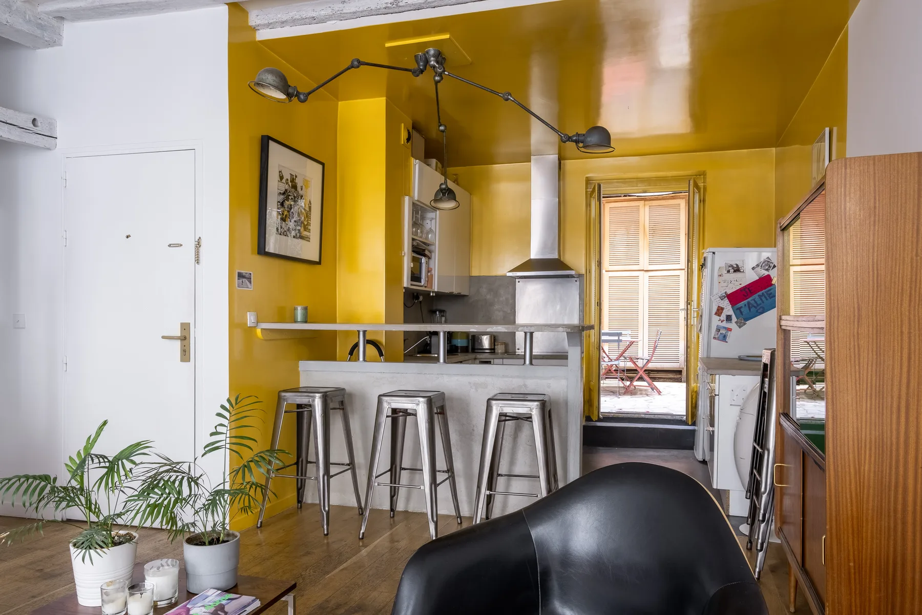 A yellow-walled kitchen with a counter, stools, and plants near the entrance.