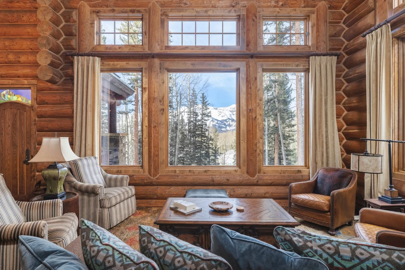 A log-cabin living room with seating arranged around a low wooden table and large windows.