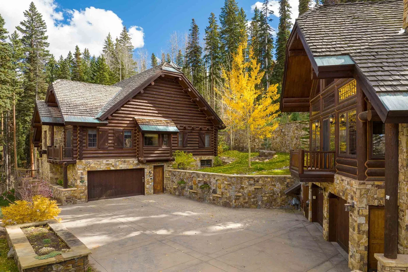 Log cabin-style homes with stone lower walls sit beside a paved driveway among trees.