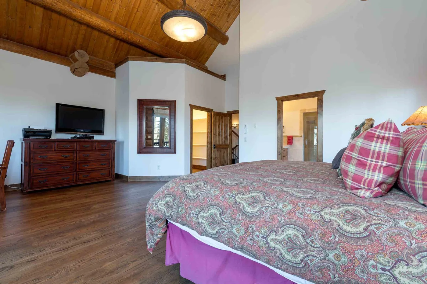 A bedroom with a patterned bed, wooden dresser, and television beneath a wooden ceiling.