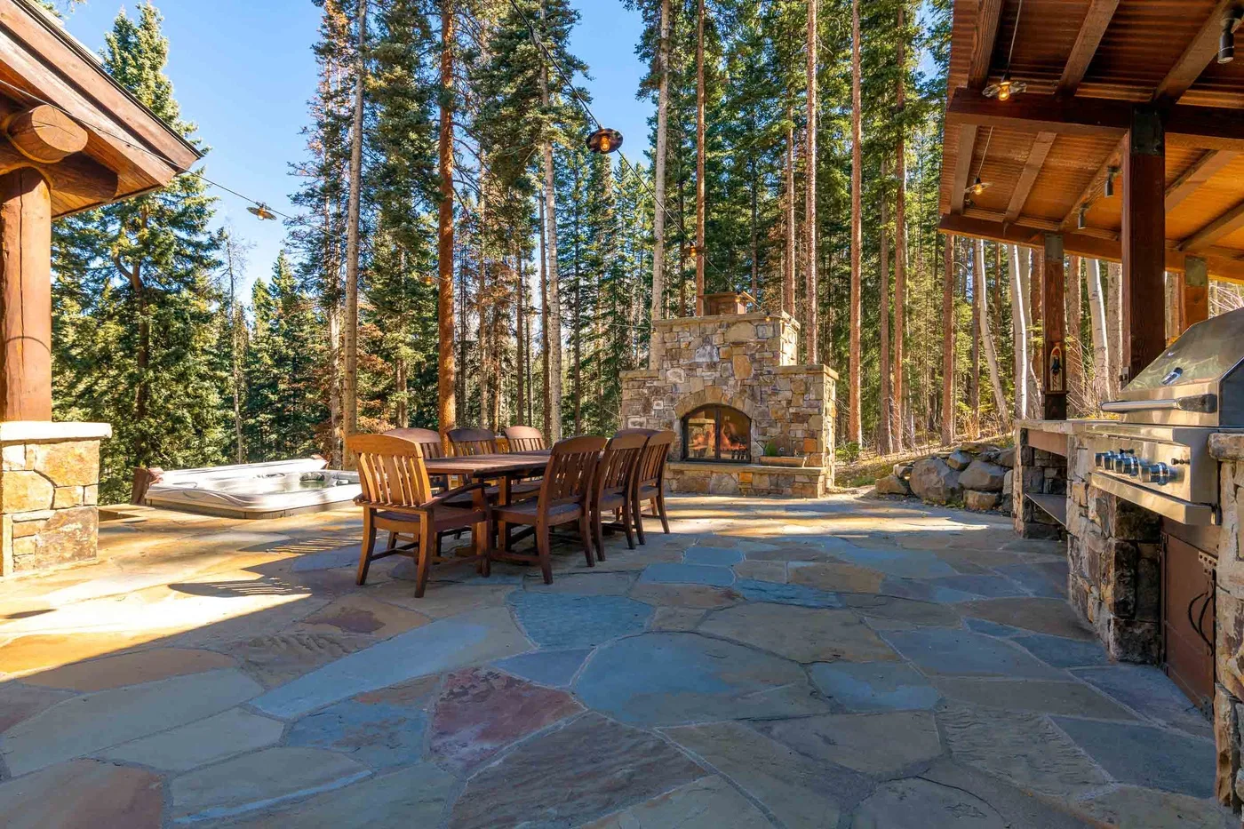 An outdoor patio with a dining table, stone fireplace, and surrounding pine trees.