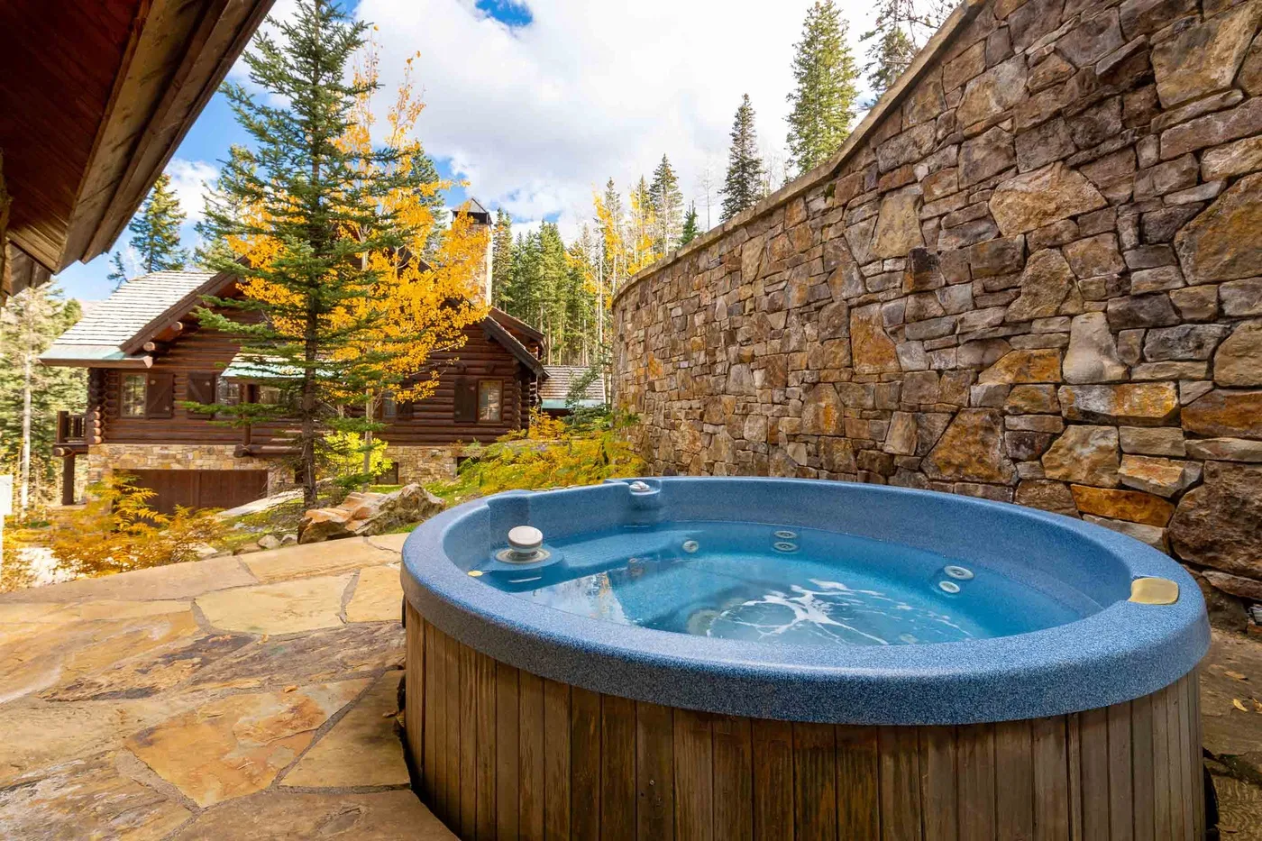An outdoor hot tub sits beside a stone wall, with a wooded cabin in the background.