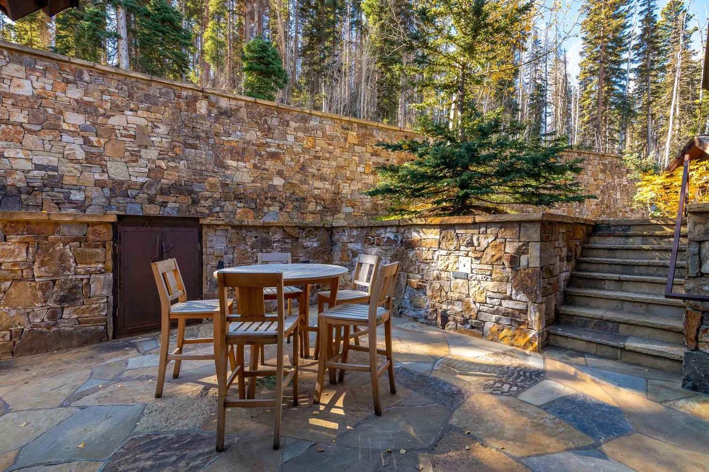 A stone patio with outdoor seating sits beside a wooded slope and steps.
