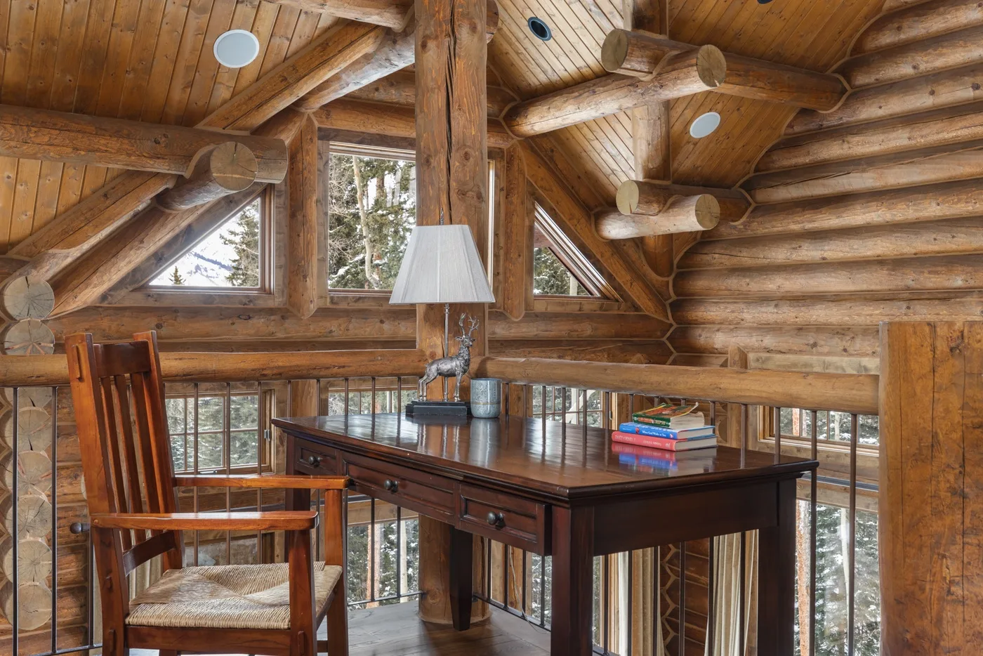 A wooden loft workspace with a desk, chair, and windows beneath a timber ceiling.