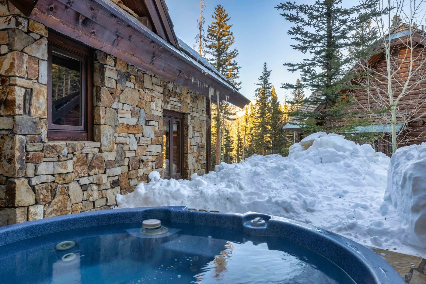 An outdoor hot tub sits beside a stone building in a snowy mountain setting.