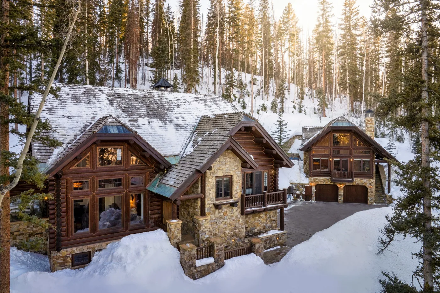 A large timber-and-stone cabin sits among snow-covered ground and pine trees.