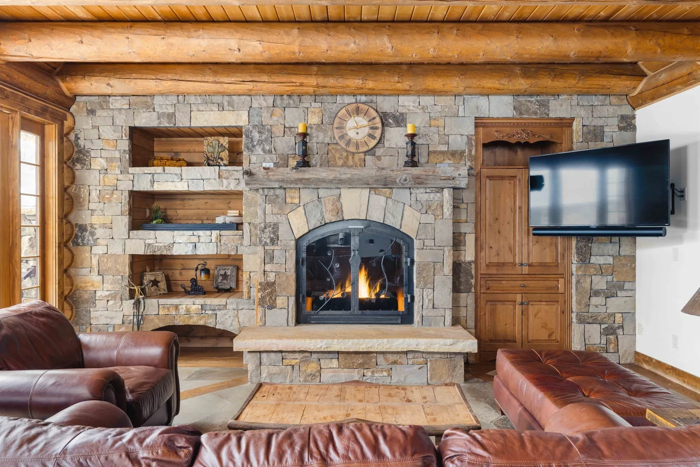 A rustic living room centers on a stone fireplace, with leather seating and a television.