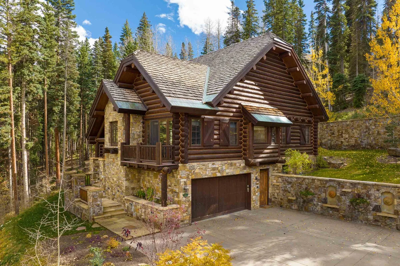 A log cabin with a stone base stands among tall trees.