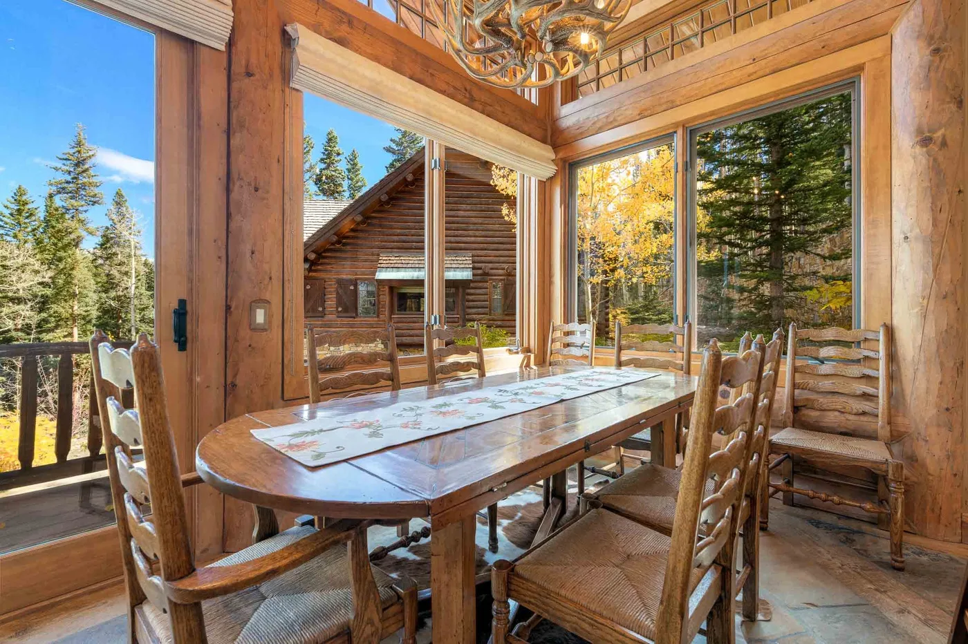 A bright dining area with a long wooden table beside large windows overlooking trees.