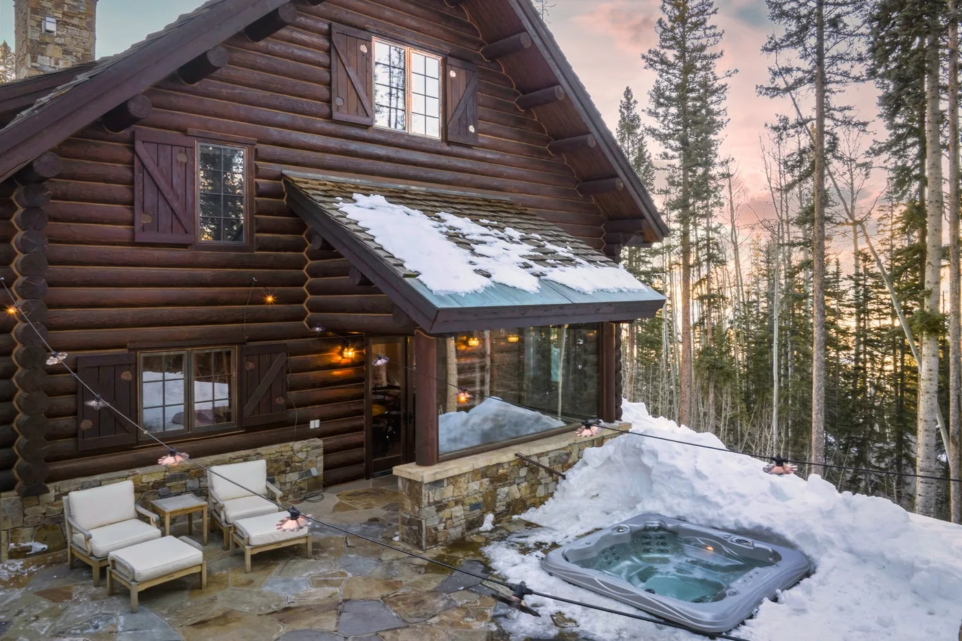 A log cabin with an outdoor hot tub sits among snow-covered trees.