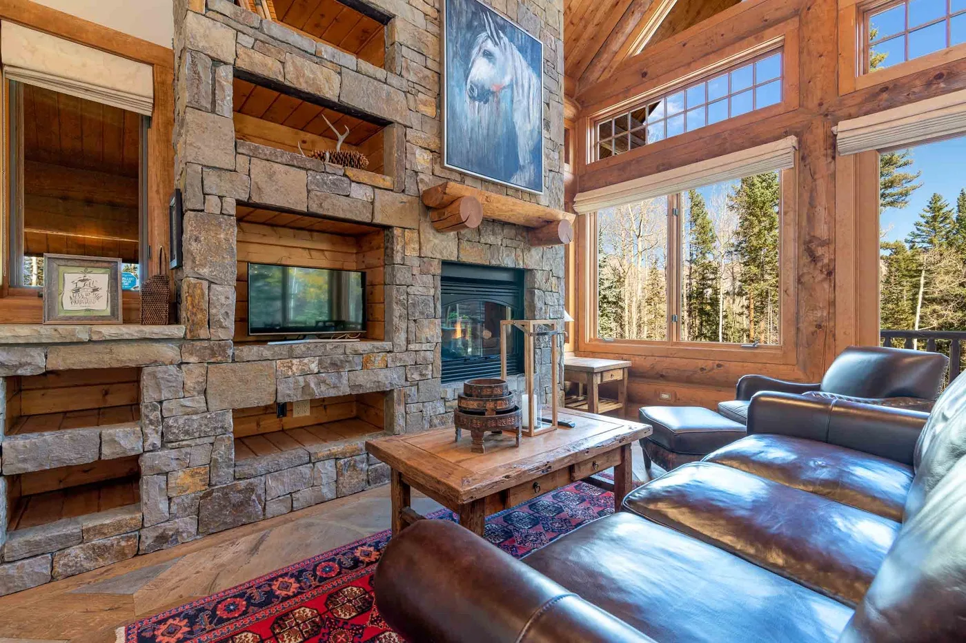 A rustic living room with a stone fireplace, wooden table, and large windows.