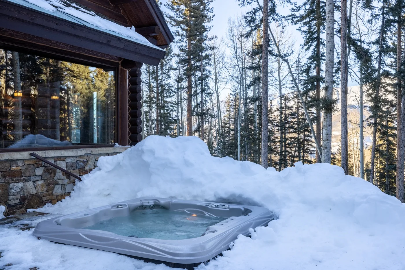 A hot tub sits in deep snow beside a log cabin among pine trees.