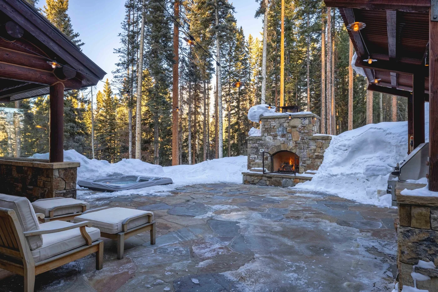 A stone patio with outdoor seating and a lit stone fireplace, surrounded by snow and trees.