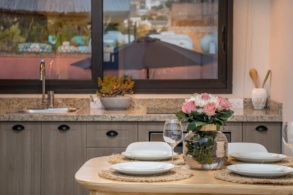 A dining table with plates and flowers sits beside a kitchen counter and sink.