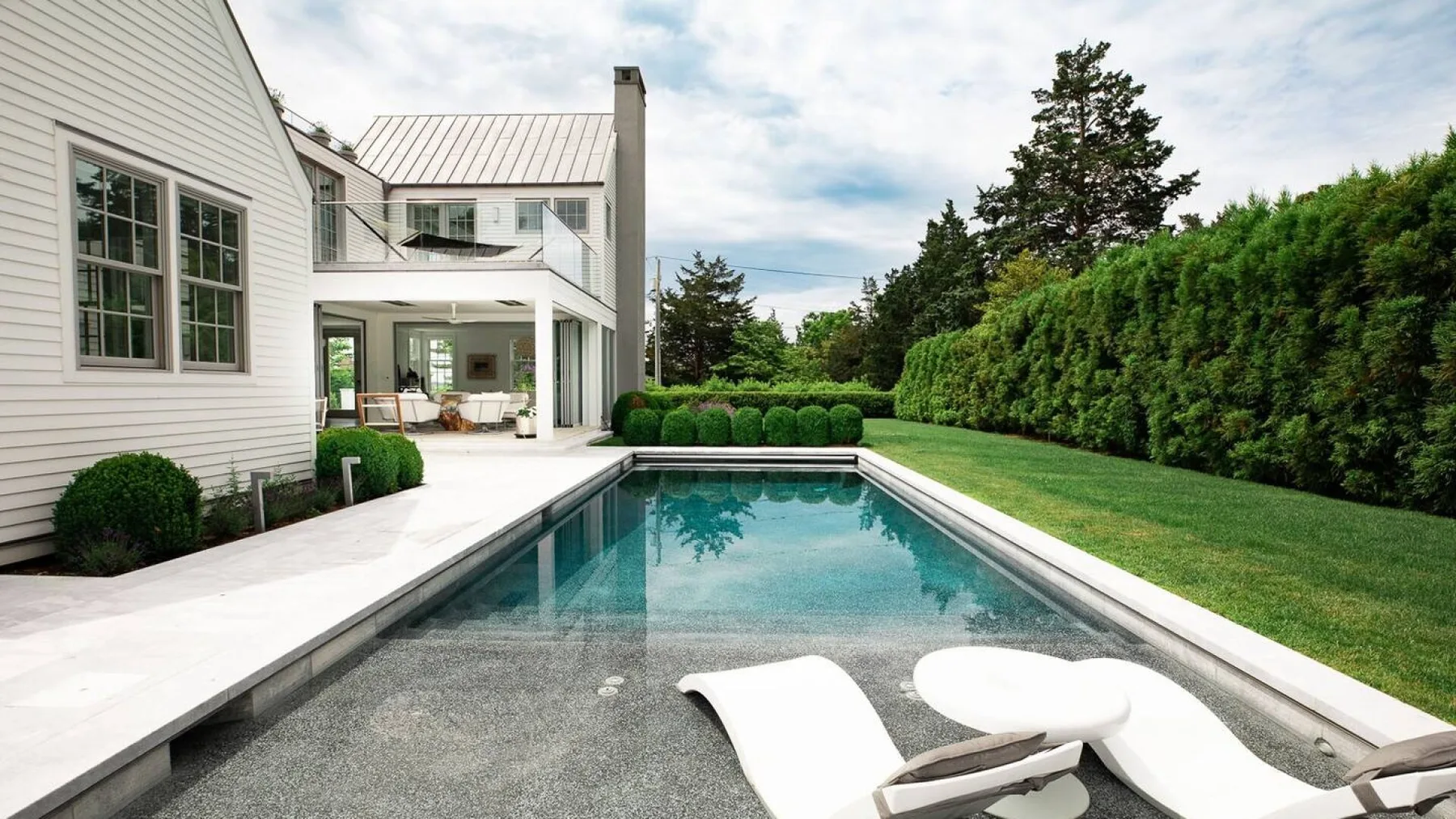 Una piscina rectangular al aire libre está junto a una casa blanca y un césped verde.