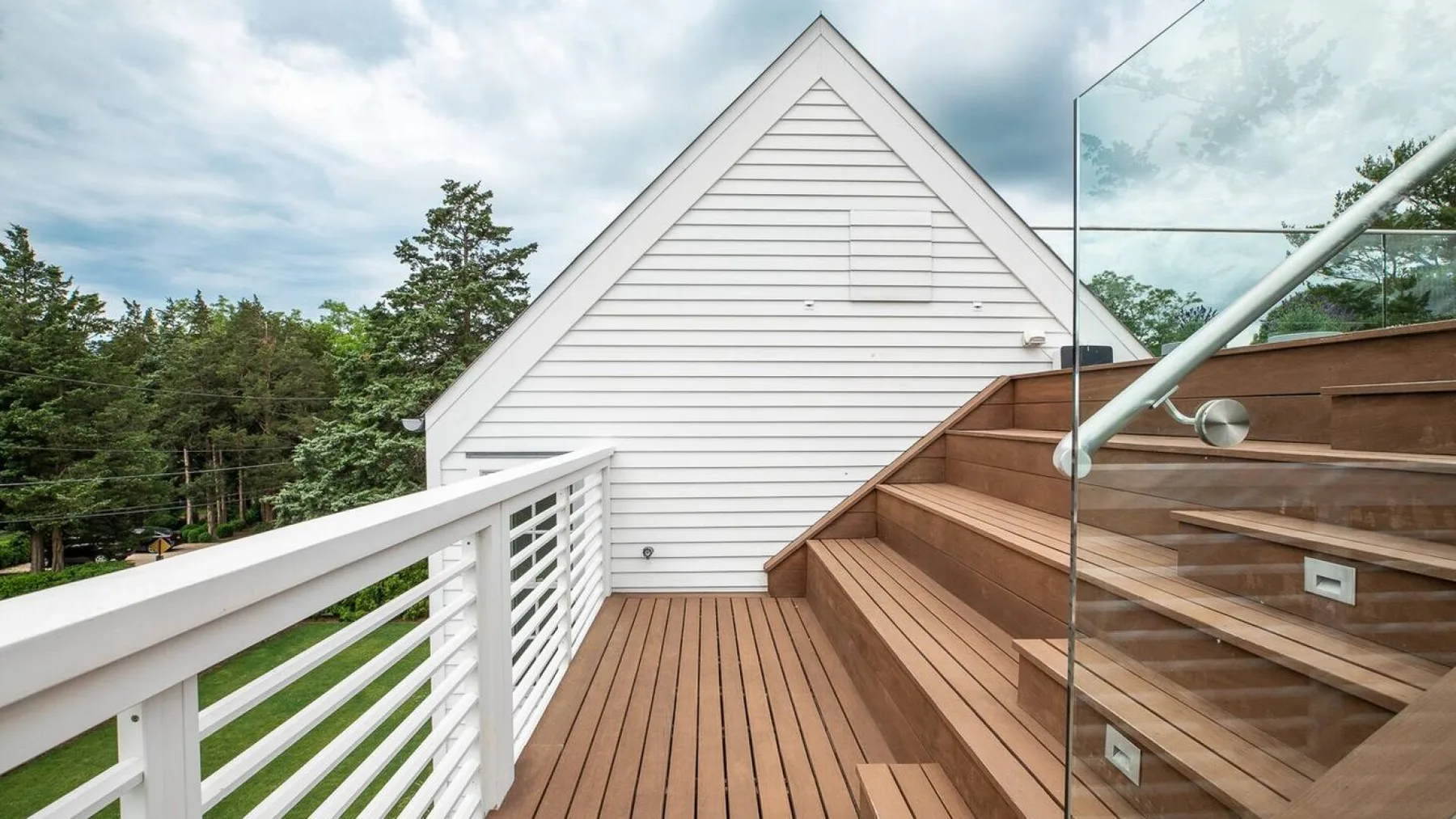 Una terraza exterior de madera junto a una pared blanca a dos aguas, con barandilla de vidrio.