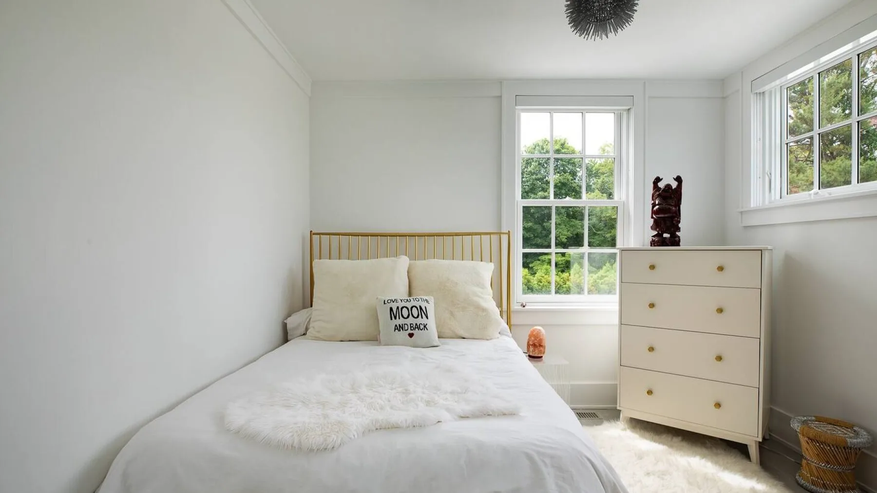 Un dormitorio luminoso con una cama bajo las ventanas y una cómoda al lado.