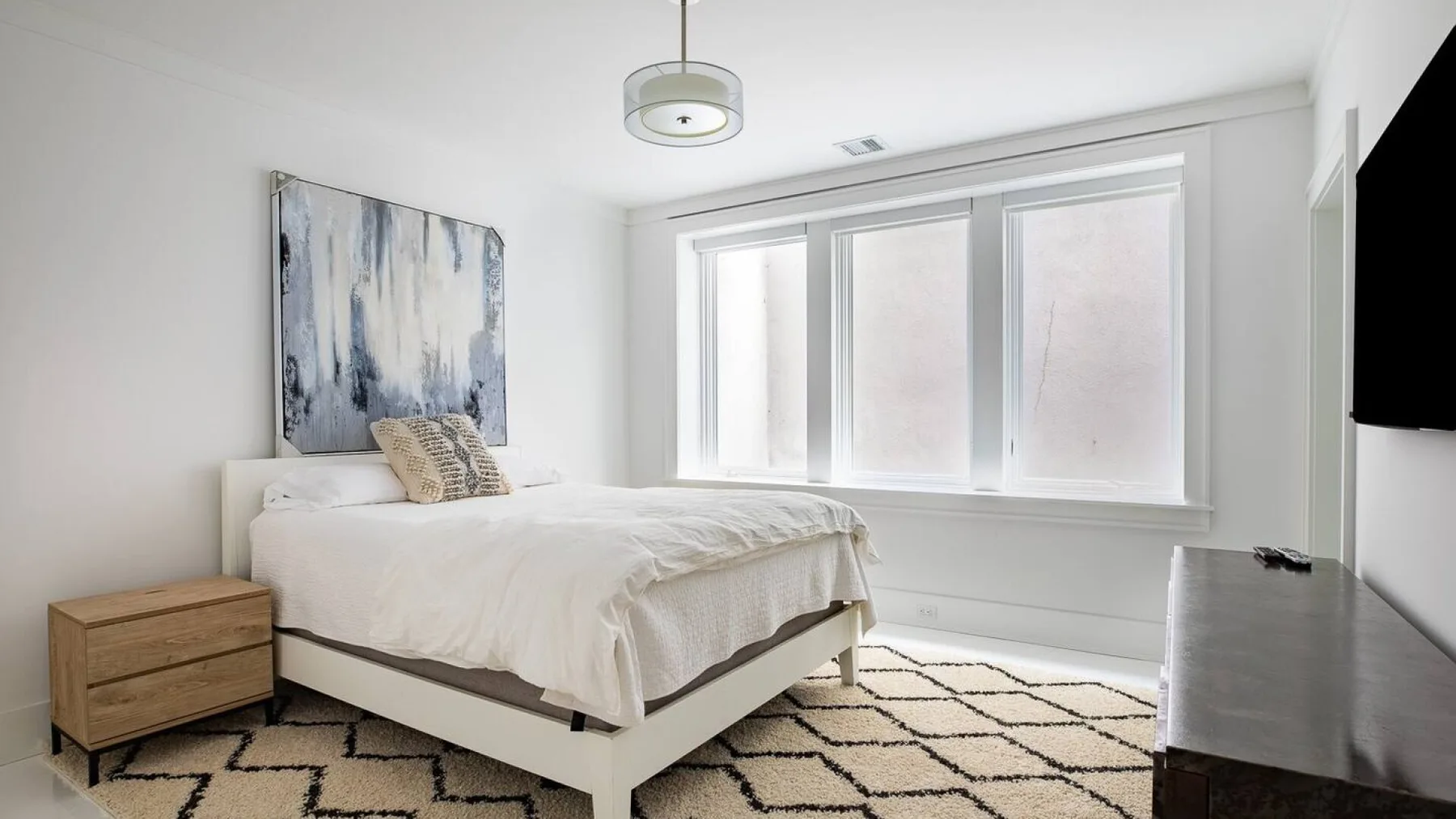 Un dormitorio luminoso con una cama tendida, ventanas grandes y una alfombra estampada.