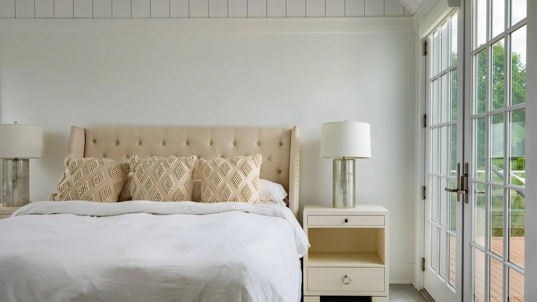 Un dormitorio luminoso con una cama, cojines estampados y ventanas altas.