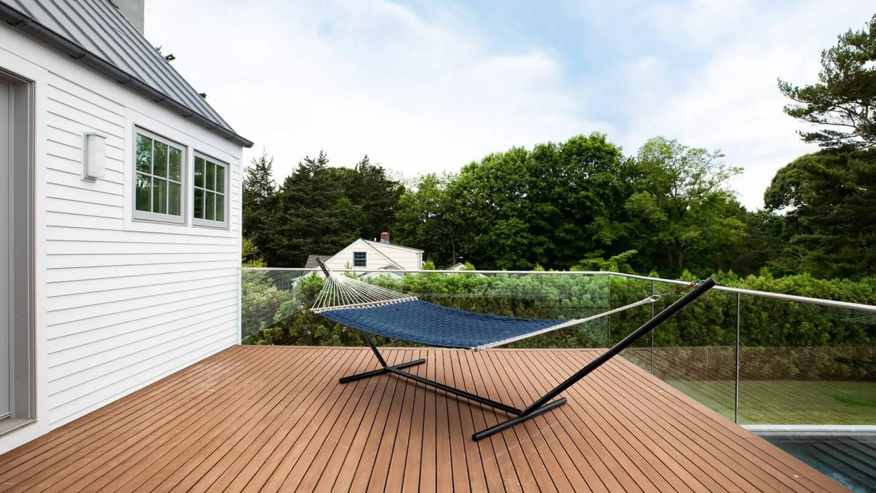 Una terraza de madera junto a una casa blanca tiene una hamaca y una barandilla de vidrio.