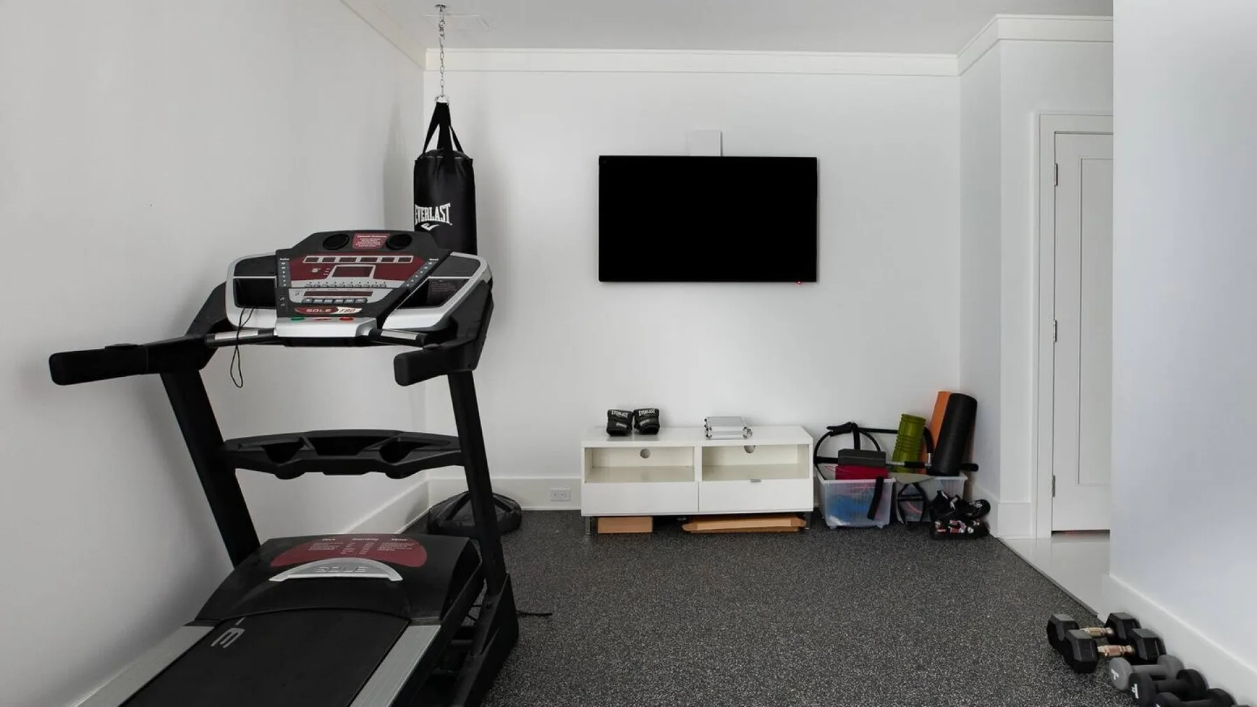 Zona de ejercicio en casa con una cinta de correr, un saco de boxeo y un televisor en la pared.