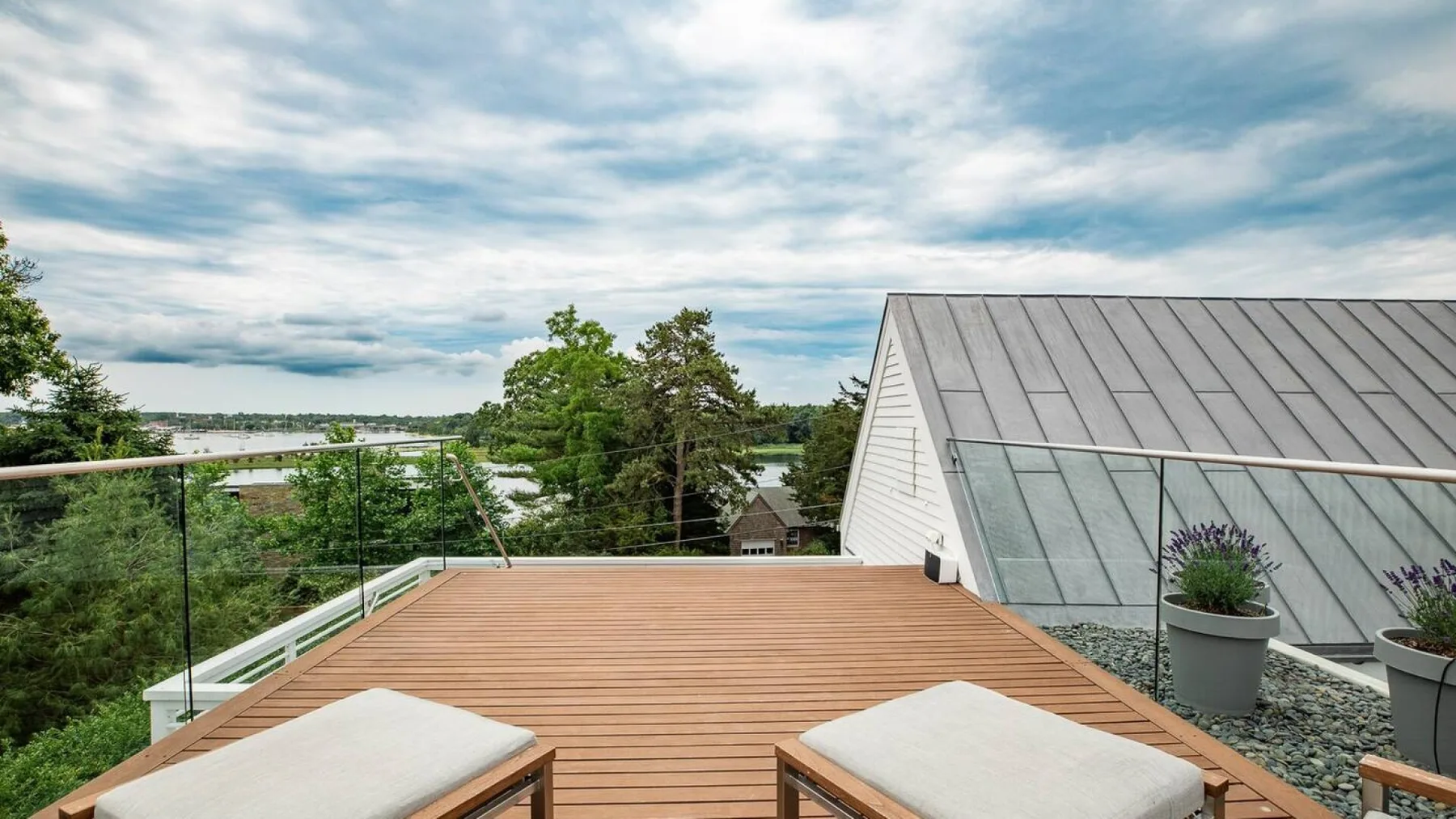 Una terraza de madera ofrece vistas a árboles y agua junto a un techo inclinado de vidrio.