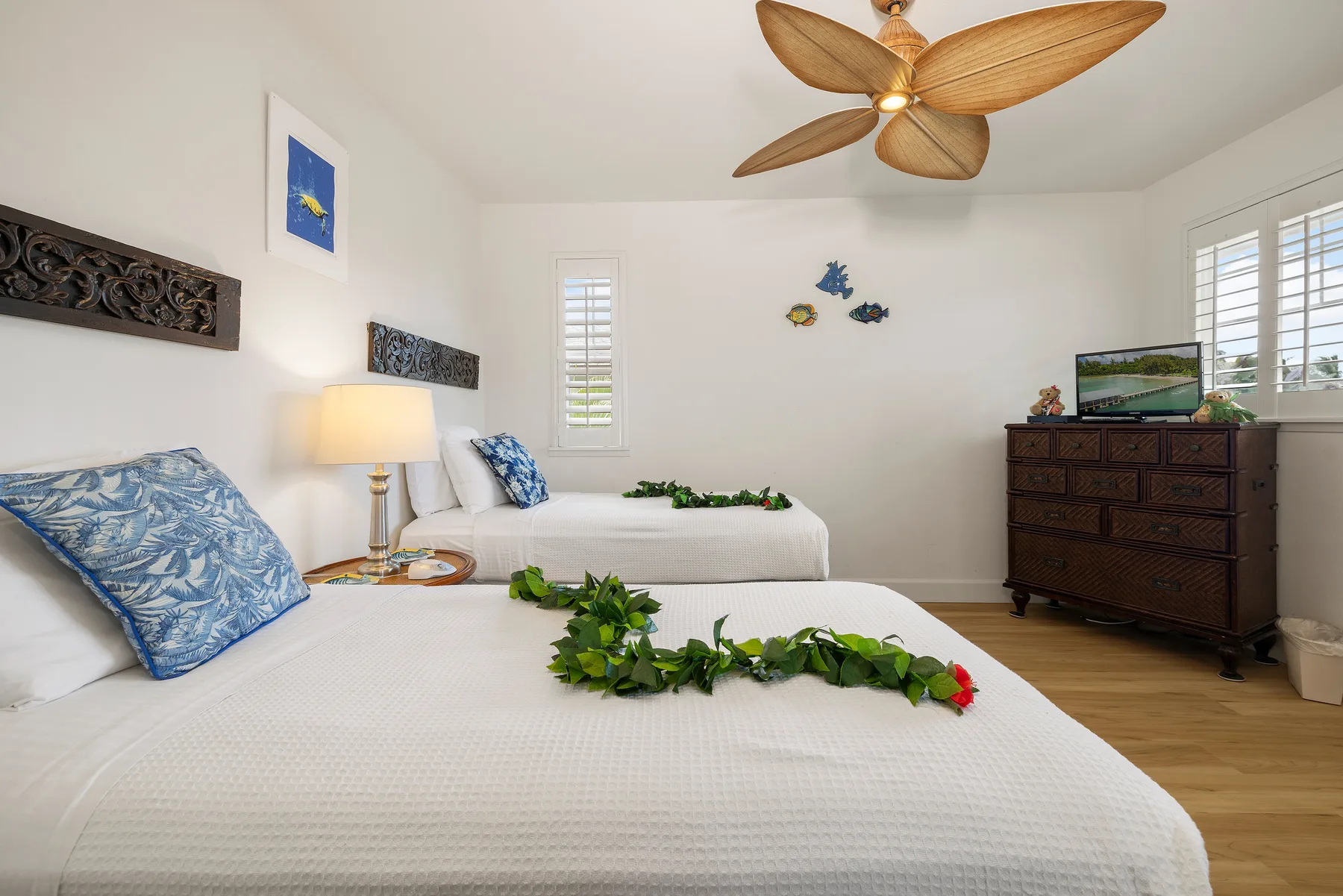 A bedroom with two beds, floral greenery, and a wooden ceiling fan.